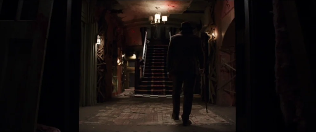 Abattoir (2016) movie still: fullbody — A dimly lit foyer features a central staircase with a patterned carpet. A man in a dark hat and suit walks…
