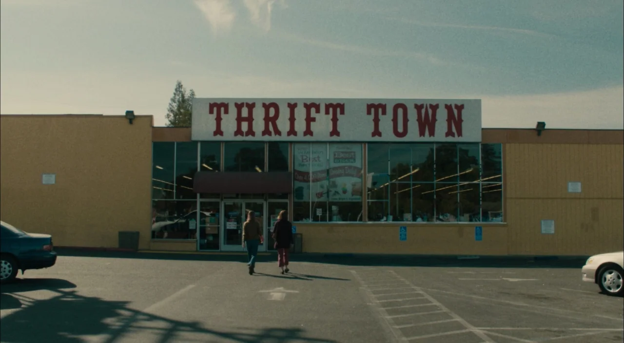 Lady Bird (2017) movie still: wide — A low-slung, yellow-walled retail building features large glass windows and a prominent red sign. Two young…