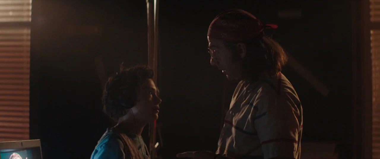 Honey Boy (2019) movie still: medium — A dimly lit room features horizontal blinds filtering dim light against a dark background. An adult man with…