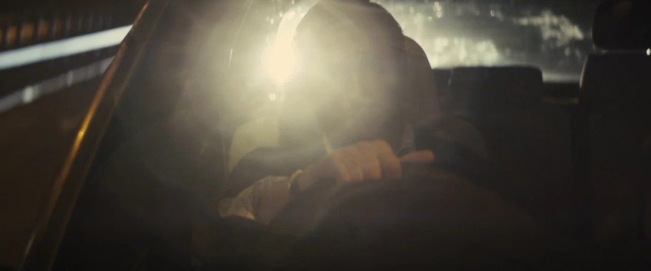 Locke (2013) movie still: medium — A dark car interior at night is lit by a piercing light source. A man grips the steering wheel while driving…