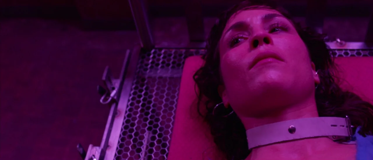Rupture (2016) movie still: closeup — A woman lies on a metallic mesh surface with a heavy white collar fixed around her neck. Bright magenta…