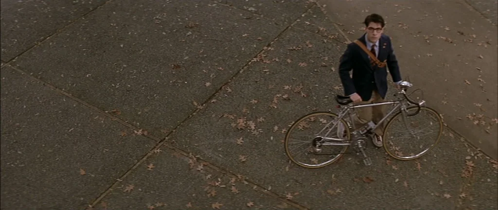 Rushmore (1998) movie still: fullbody — A paved area is covered in scattered dry brown autumn leaves. A young man wearing a blazer and spectacles…