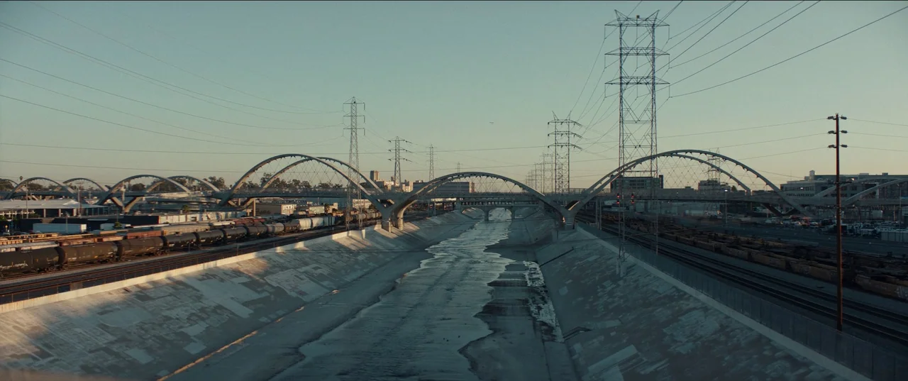 The Greatest Hits (2024) movie still: establishing — A concrete drainage channel in an urban industrial zone with parallel rail tracks. Multiple…