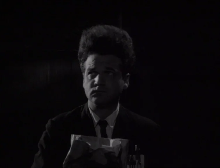 Eraserhead (1977) movie still: medium — A man stands in an unidentifiable, pitch-black interior space. He wears a dark suit and maintains a…
