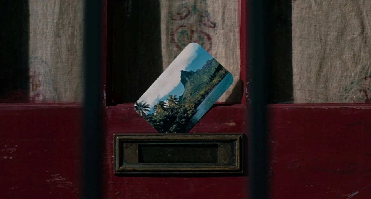 Inherent Vice (2014) movie still: closeup — A close-up view of a deep red wooden front door with a metal slot. A postcard showing a lush tropical…