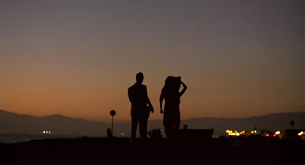 Morvern Callar (2002) movie still: wide — A silhouette of a man and a woman stand against a twilight sky near water. Distant city lights appear…