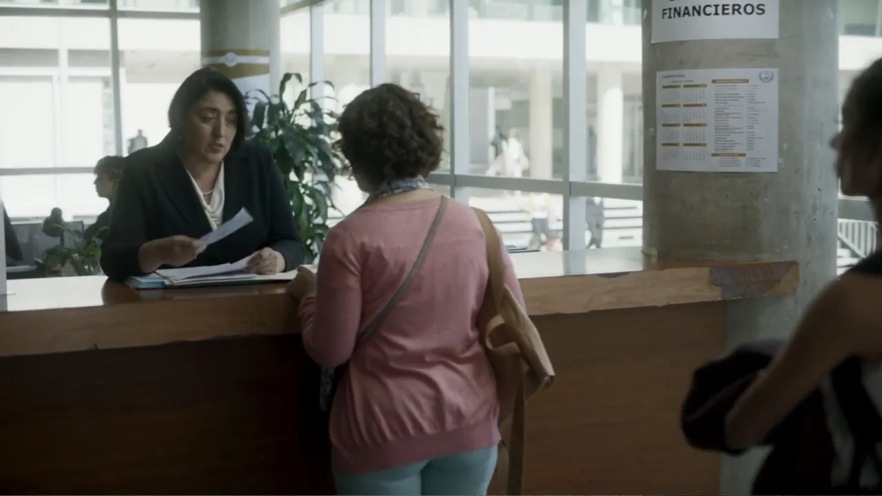 Lecciones para canallas (2022) movie still: medium — A bank interior features a wooden counter in front of large windows. A teller assists a…