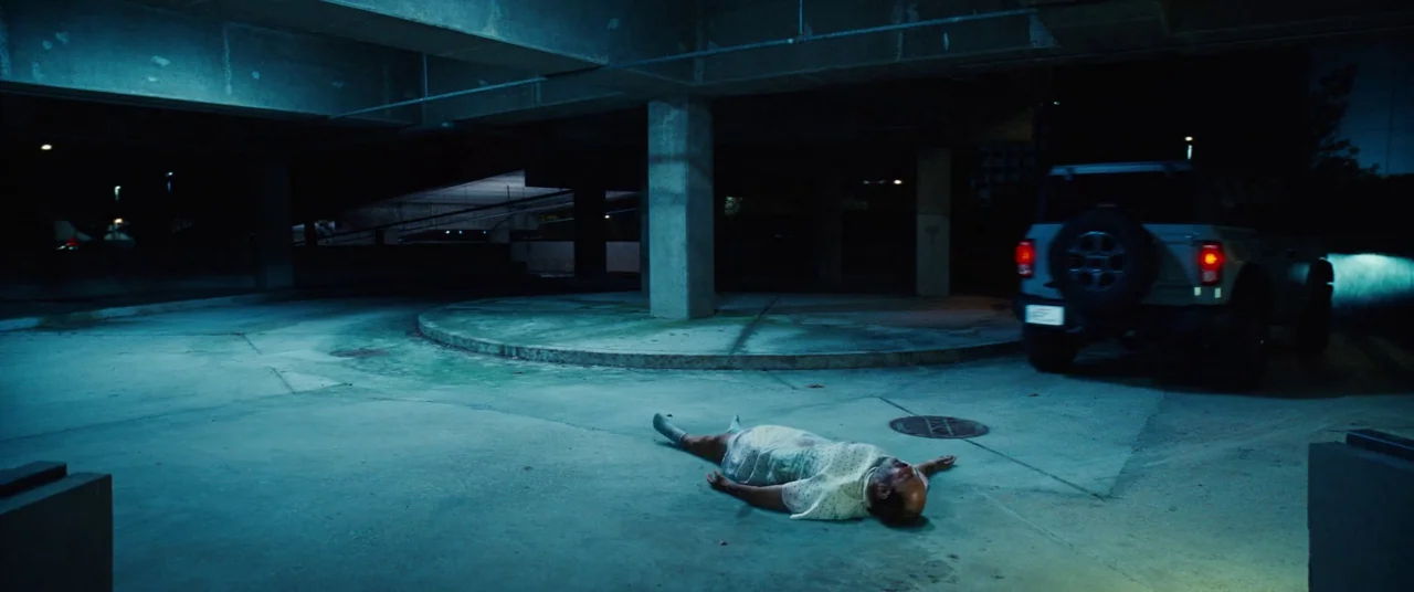 Kinds of Kindness (2024) movie still: wide — A concrete parking garage level is illuminated by cold artificial light at night. A man wearing a…