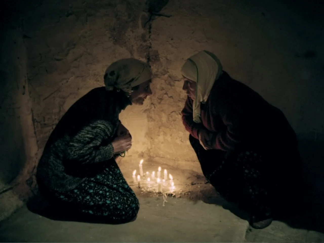 Don't Let Them Shoot the Kite (1989) movie still: medium — A dimly lit room with rough stone walls surrounds a cluster of lit candles. Two women…
