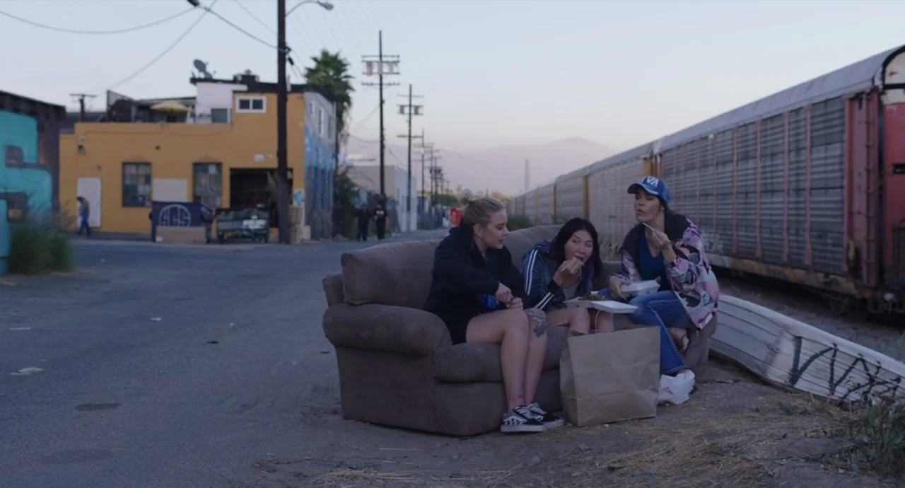 Pleasure (2021) movie still: fullbody — An abandoned sofa sits on a dirt patch near railroad tracks in an industrial area. Three young women sit…