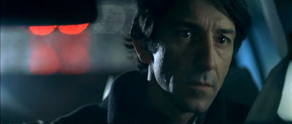 Mortal Transfer (2001) movie still: closeup — The interior of a car at night with a man driving in darkness. Two glowing red blurred taillights are…