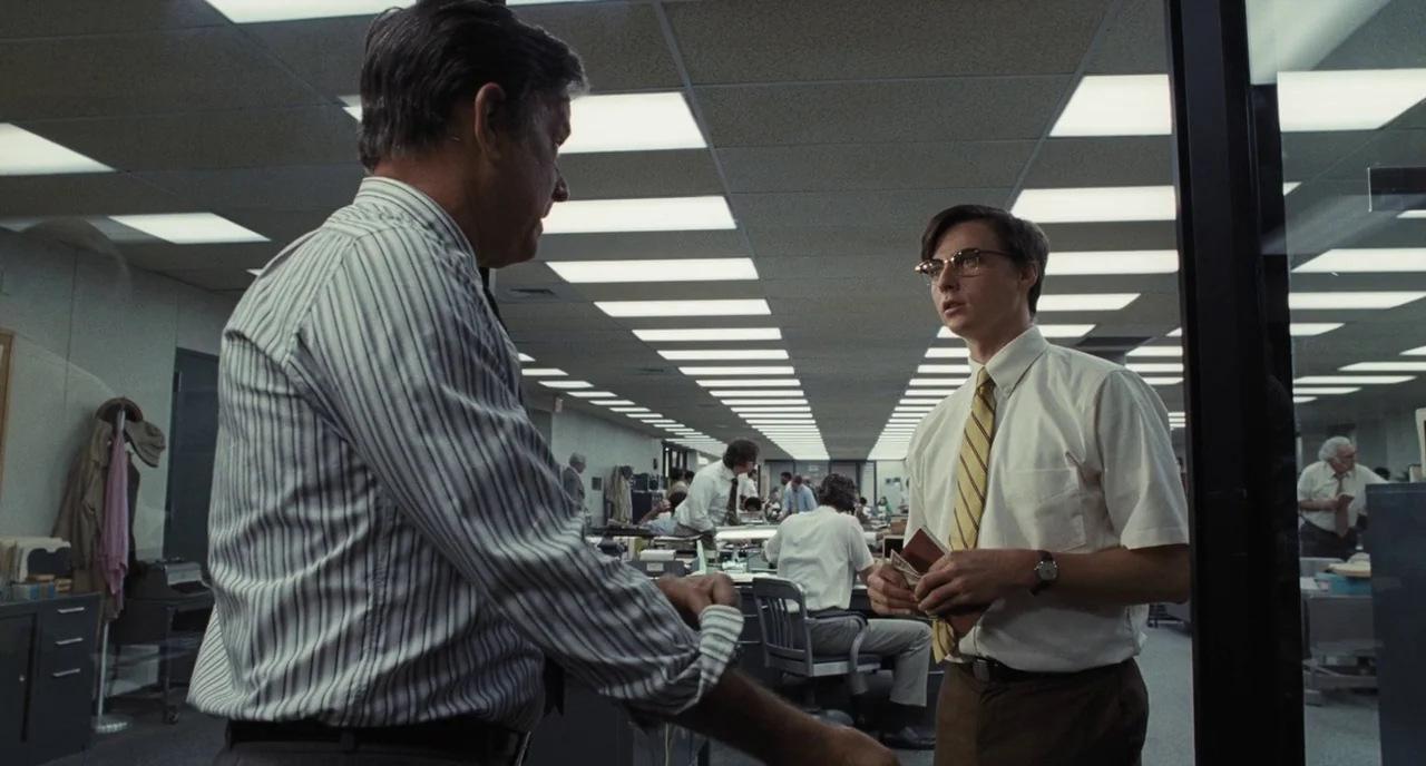 The Post (2017) movie still: medium — An open-plan corporate space features rows of desks under repetitive fluorescent ceiling light panels. Two men…