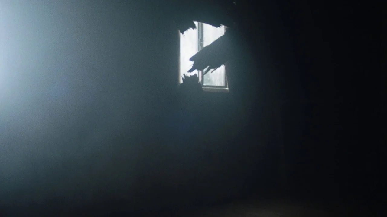 Snowbound (2017) movie still: establishing — A dark, dilapidated interior space features a small window blocked by jagged, broken wall debris.…