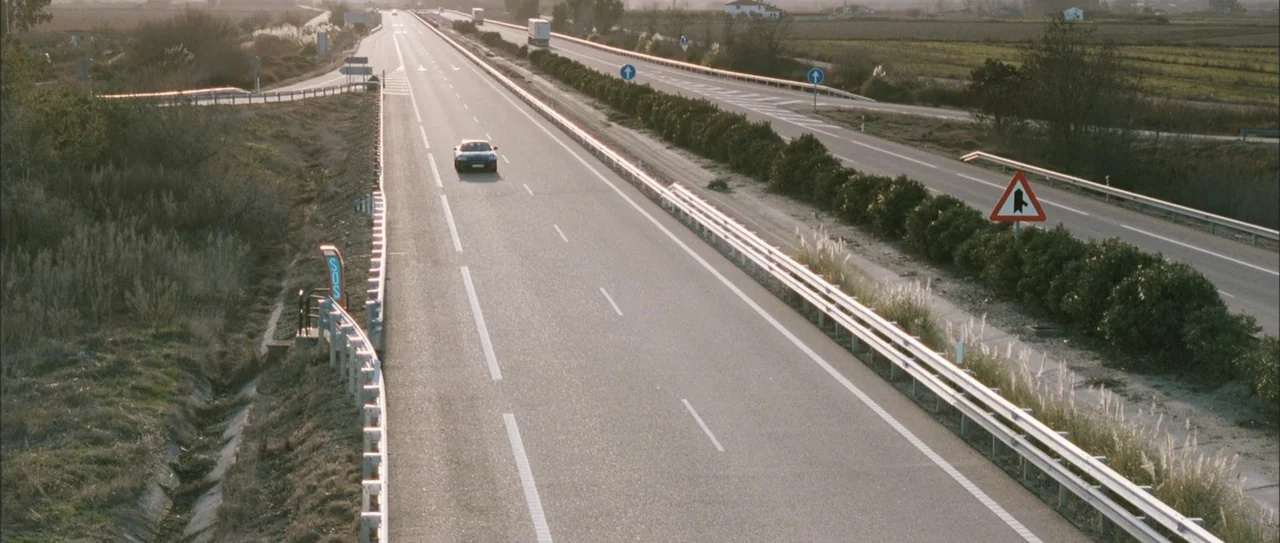 Rivals (1972) movie still: establishing — An asphalt highway stretches into the distance through a flat, rural landscape. A solitary car drives…