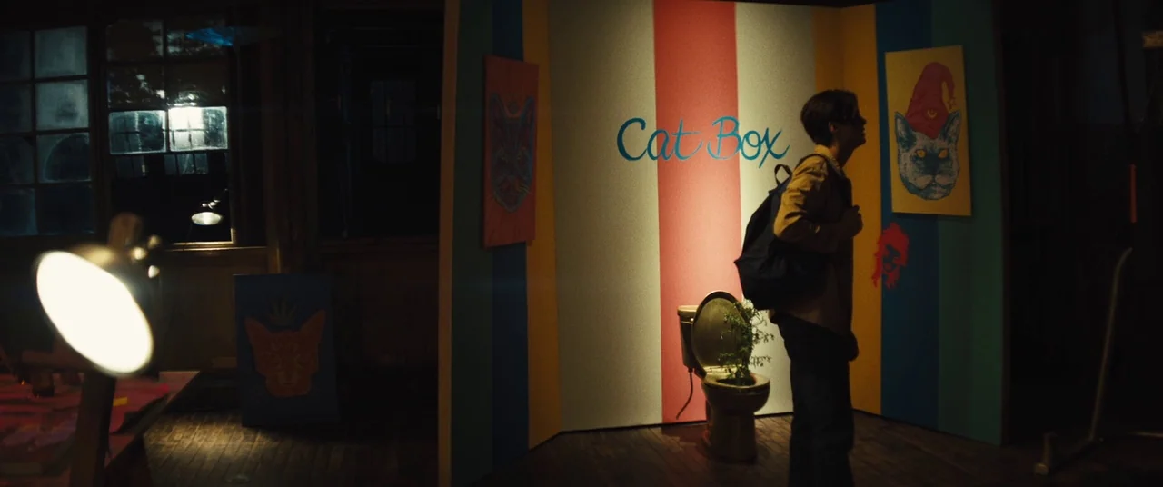 Daniel Isn’t Real movie still: fullbody — A dimly lit art space features colorful panels displaying cat-themed illustrations on the walls. A young…
