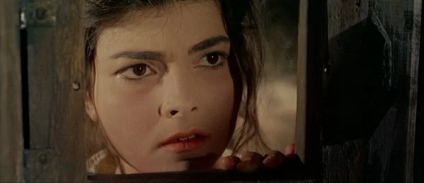 Django Kill (1967) movie still: closeup — A woman looks through a rectangular opening in a dark, weathered wooden partition. Her face fills the…