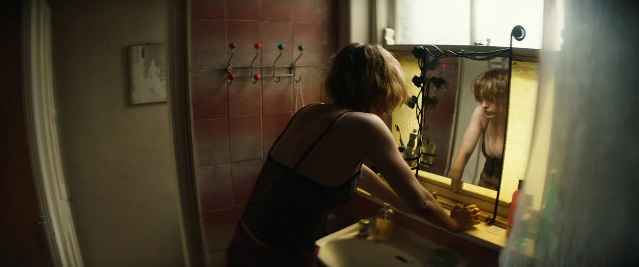 The Rhythm Section (2020) movie still: medium — A small bathroom features a tiled red wall with a multi-colored coat rack. A young woman leans over…