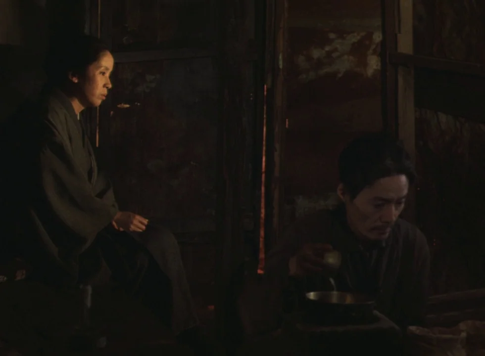 Dodes’ka-den (1970) movie still: medium — A dimly lit, cramped wooden interior contains two people in traditional robes. The man holds a small metal…