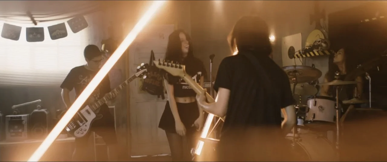 Death to Summer (2021) movie still: medium — A cluttered room used for band practice features paper decorations and musical equipment. Four young…