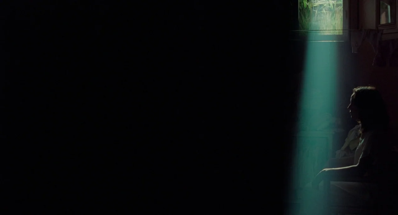 Babysitter (2022) movie still: medium — A dim room is occupied by a solitary figure near a small high window. A tight beam of daylight casts a…