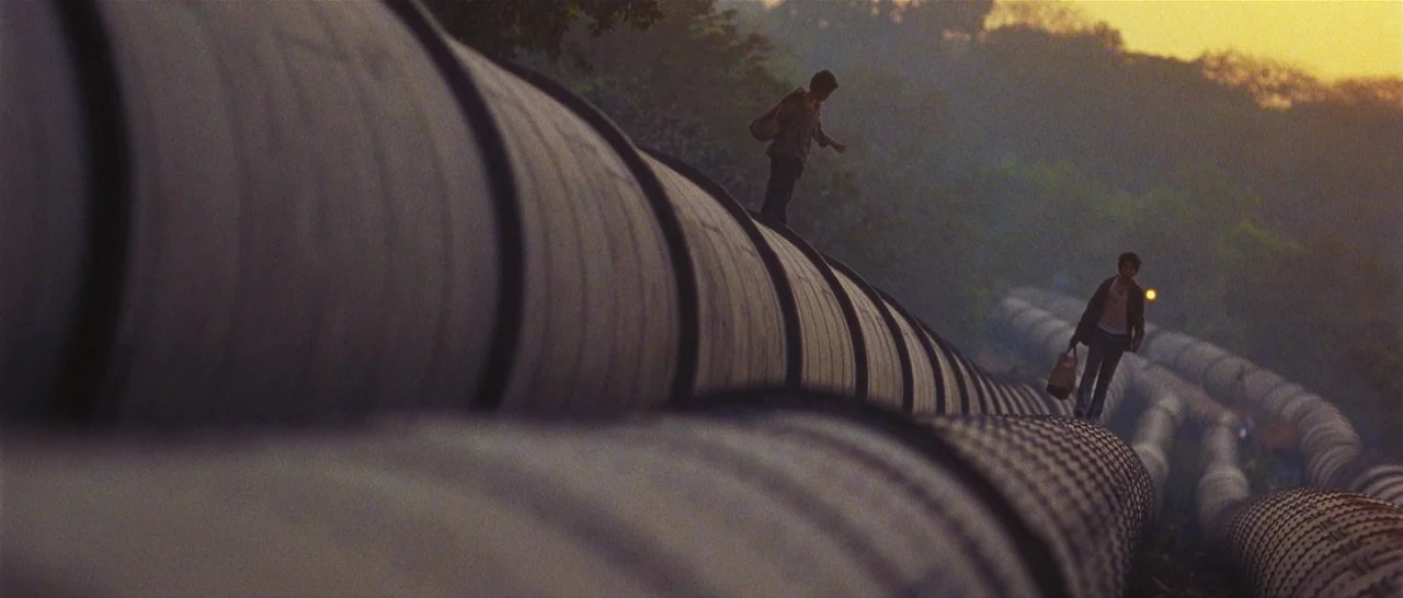 Slumdog Millionaire (2008) movie still: wide — Two parallel rows of massive concrete pipes stretch across a hazy landscape at sunset. Two boys walk…