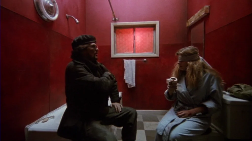 Patty Hearst (1988) movie still: wide — A windowless-style bathroom has vibrant red-painted walls and a checkered floor. A man sits on the bathtub…