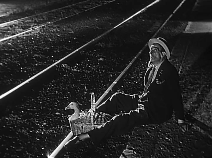 Variety Lights (1950) movie still: fullbody — A man sits on gravel beside railroad tracks at night. He is accompanied by a goose resting inside a…