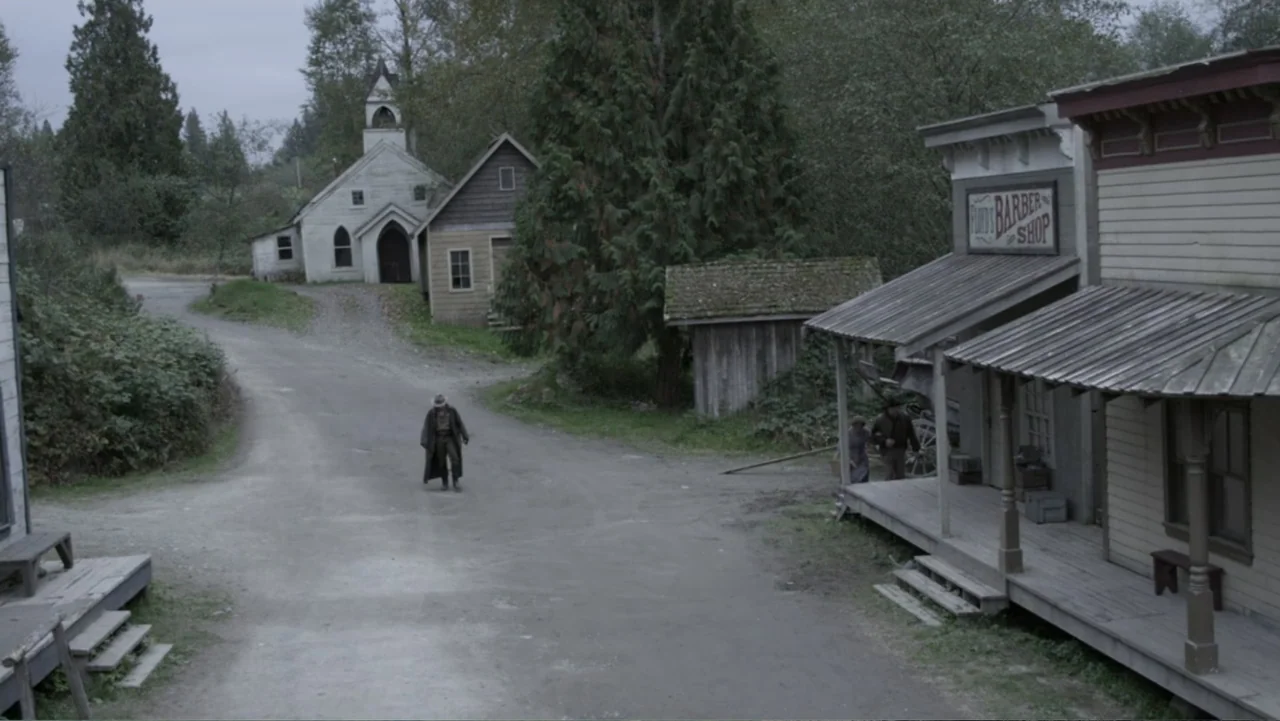 Dawn Rider (2012) movie still: wide — A rural 19th-century settlement features wooden boardwalks, a barber shop, and a white church. A man in a…