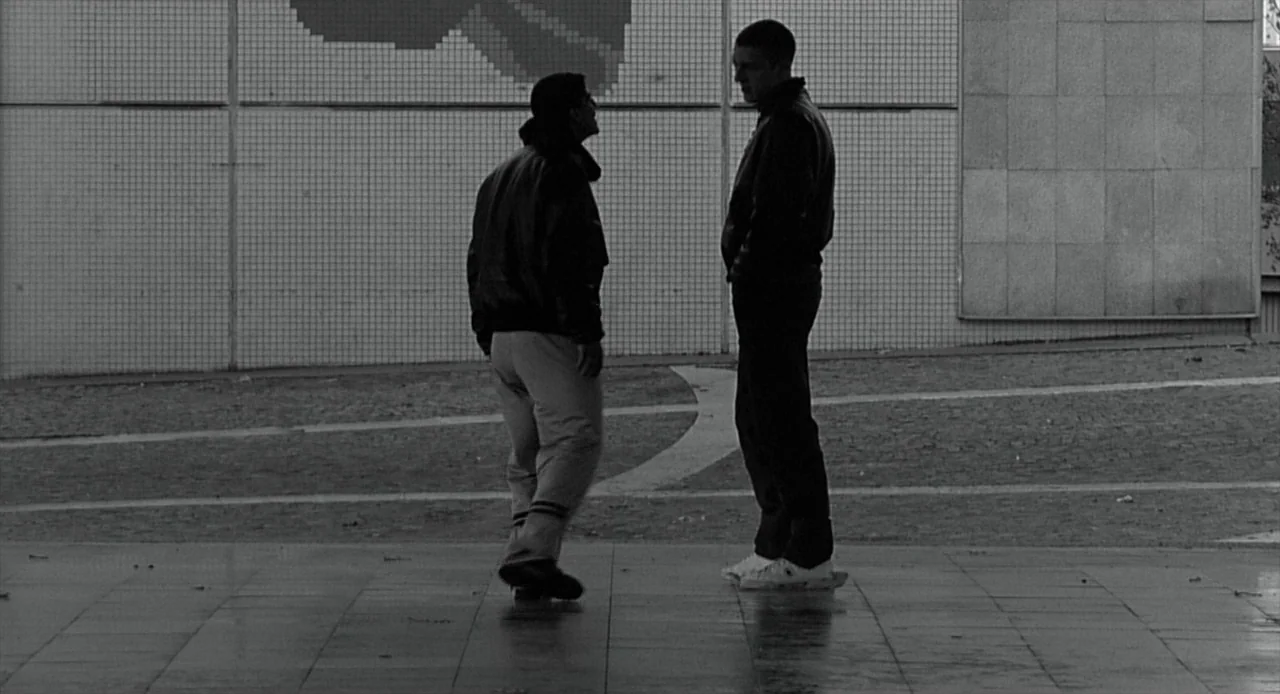 La Haine (1995) movie still: fullbody — A public square with a tiled wall and patterned concrete flooring. Two men stand facing each other in…
