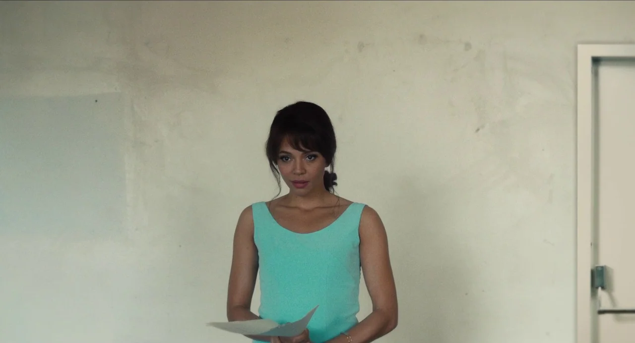 Born To Be Blue (2015) movie still: medium — A woman stands before a plain off-white wall inside an office space. She holds a document in her hands…