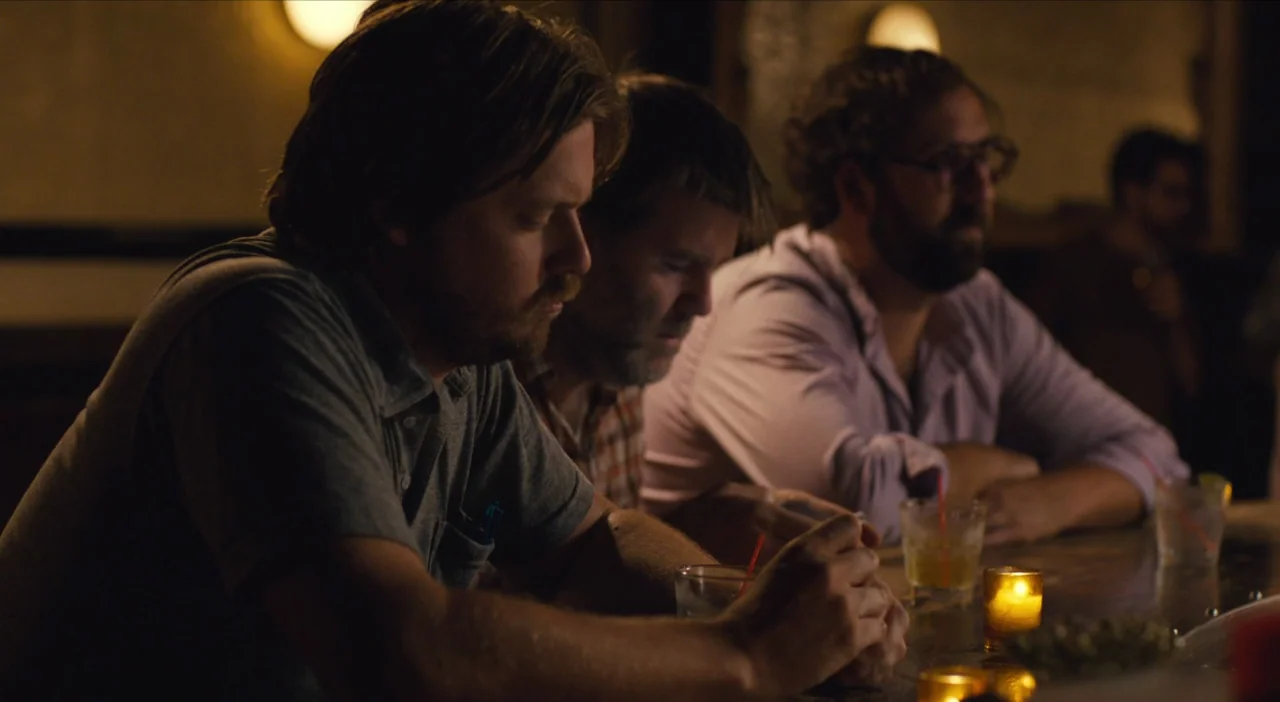 The Comedy (2012) movie still: medium — Three men sit along a wooden counter in a dimly lit, narrow space. Each man rests their arms on the surface…