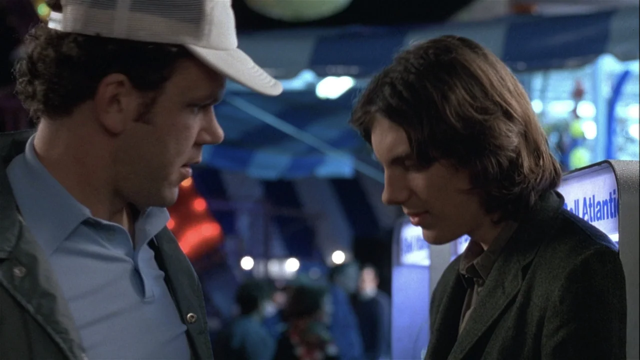 Boys (2021) movie still: medium — Two men converse at night near a glowing public telephone sign at a fairground. The taller man in a baseball cap…