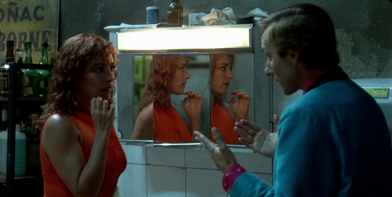 Law of Desire (1987) movie still: medium — A dimly lit restroom features white-tiled walls and a medicine cabinet mirror. A woman wearing an orange…