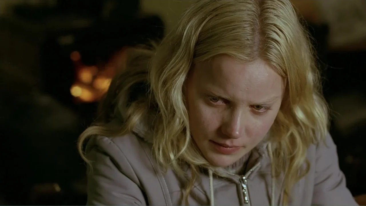 Emily (1976) movie still: closeup — A dimly lit indoor space features a flickering fire in the background. A young blonde woman in a hoodie gazes…
