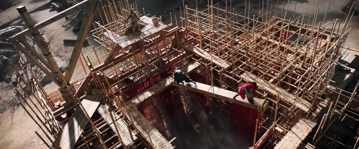 Mulan (2020) movie still: wide — A sprawling bamboo construction structure features levels of platforms and beams. Several warriors navigate the…