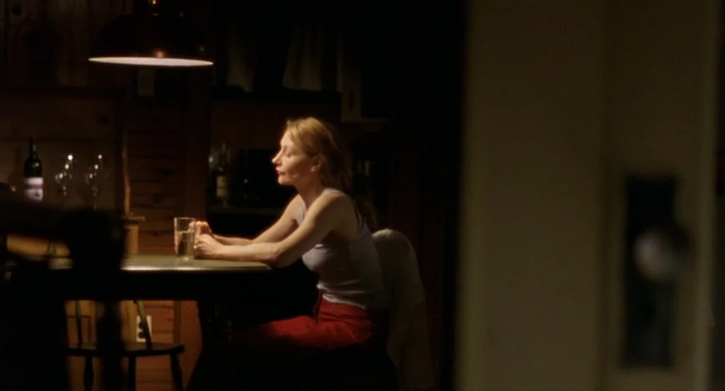 The Station Agent (2003) movie still: medium — A dark, wood-paneled room features a table set with a glass of water. A woman in a tank top sits at…
