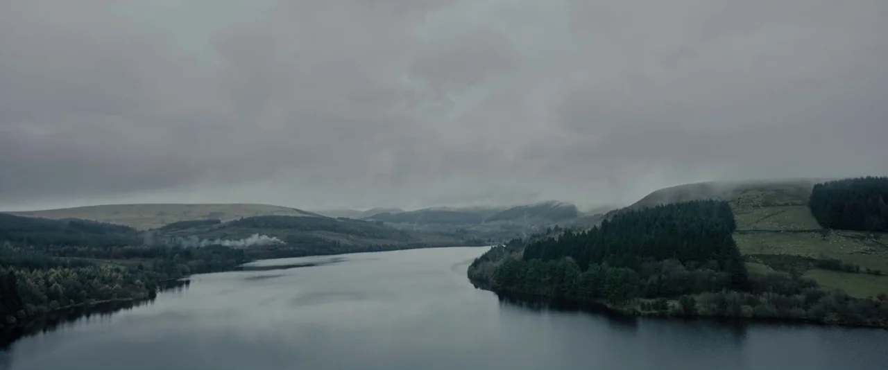 Apostle (2018) movie still: establishing — A wide, calm reservoir stretches between dark hills covered with pine trees and grass. Low-hanging grey…