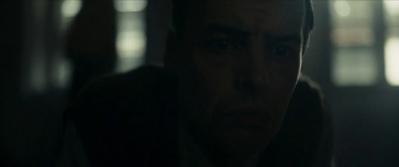 Earwig (2022) movie still: closeup — A dark, minimally lit interior with soft illumination hitting the subject's forehead and eyes. A man looks…