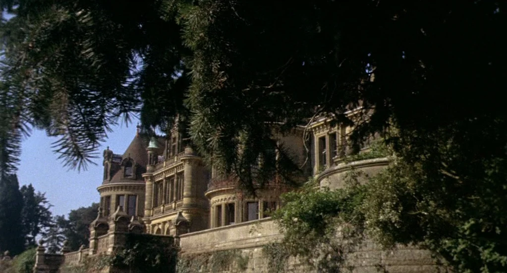 Demons of the Mind (1972) movie still: establishing — A stone mansion with turrets and intricate masonry stands behind a high wall. Dark evergreen…