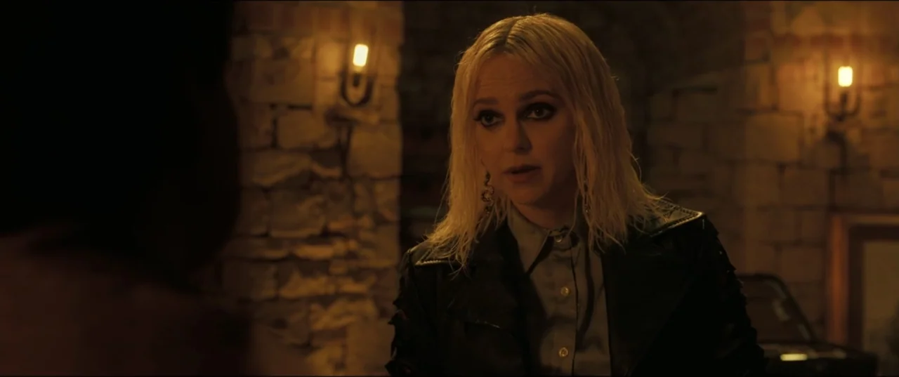 My Spy The Eternal City (2024) movie still: medium — A woman stands in a dim room with rough-hewn stone walls and lit sconces. She faces a person…