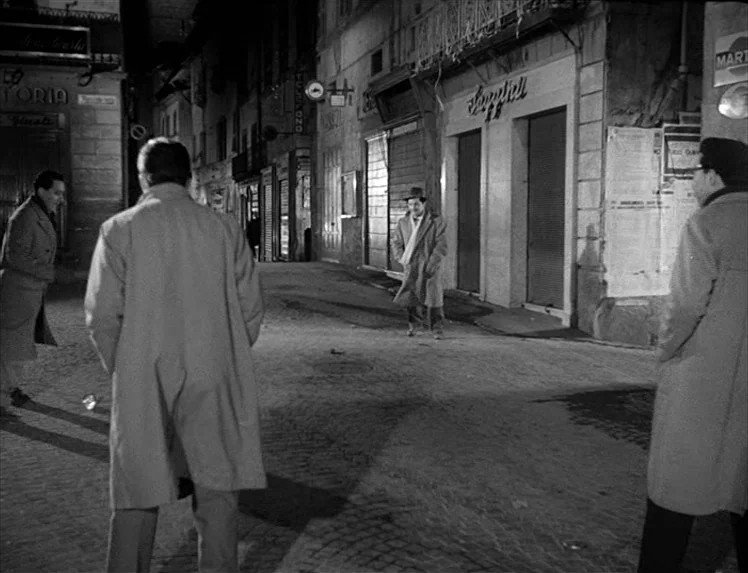 I Vitelloni (1953) movie still: fullbody — A quiet, narrow European cobblestone street at night is lined with closed storefronts. Four men in trench…