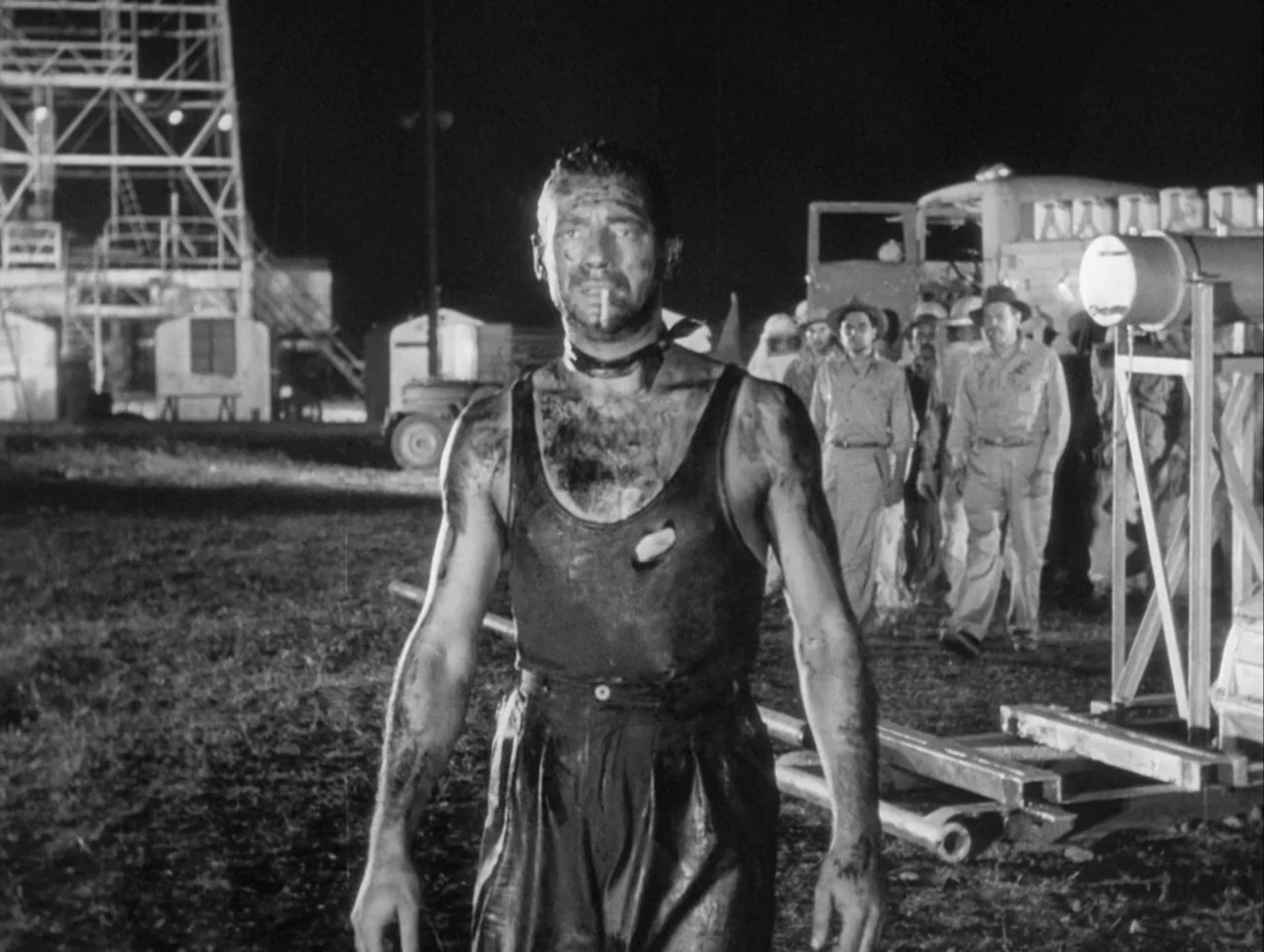 The Wages of Fear (1953) movie still: fullbody — A nighttime industrial landscape features a large oil derrick towering in the distance. A man…