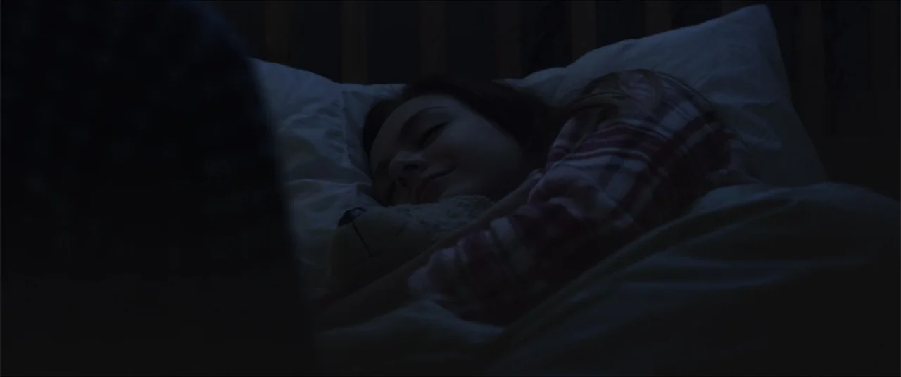 Captive (2020) movie still: closeup — A dark bedroom interior with a child sleeping under white linens. A girl rests her head on a white pillow…