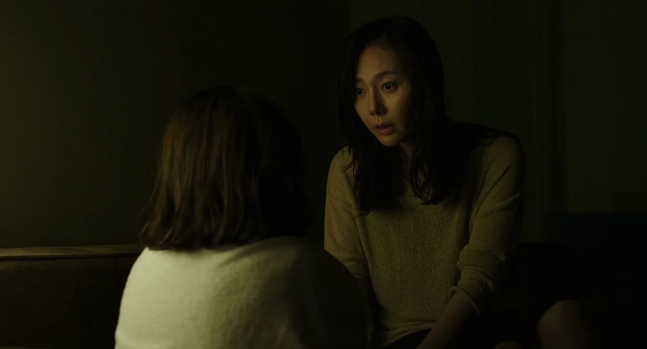 The Mimic (2017) movie still: medium — A dimly lit room with the background swallowed by dark shadows. A woman looks at the back of another person.