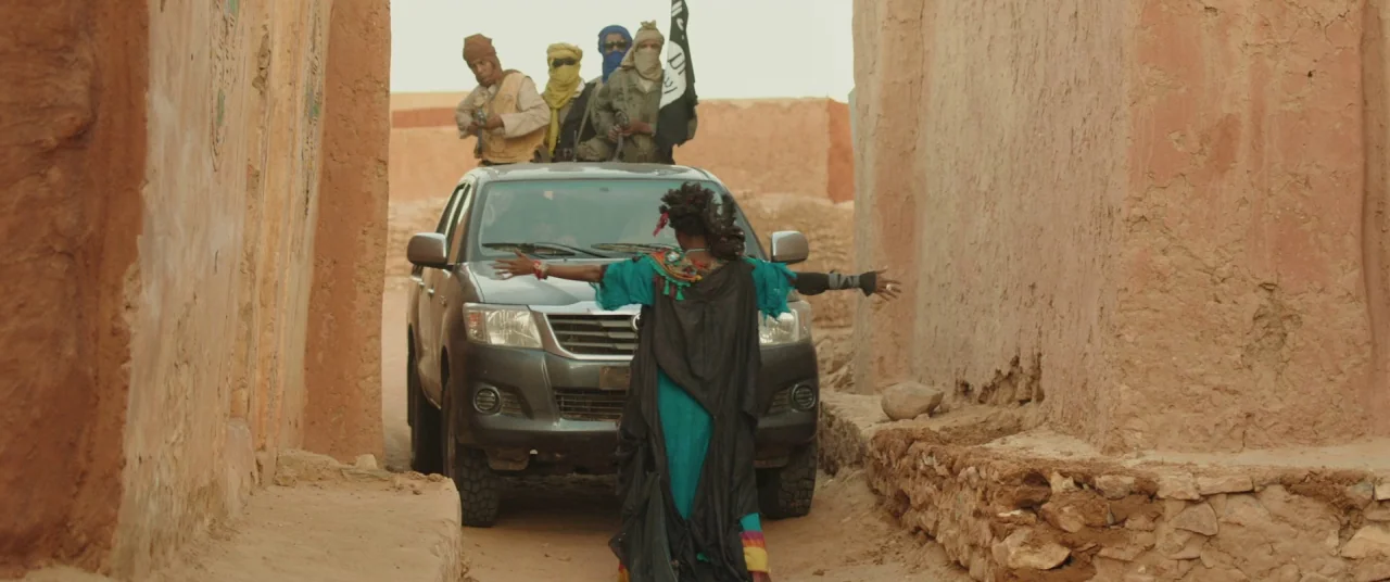 Timbuktu (2014) movie still: fullbody — A narrow alleyway features textured, sun-baked earth walls on both sides. A woman stands in the center of…