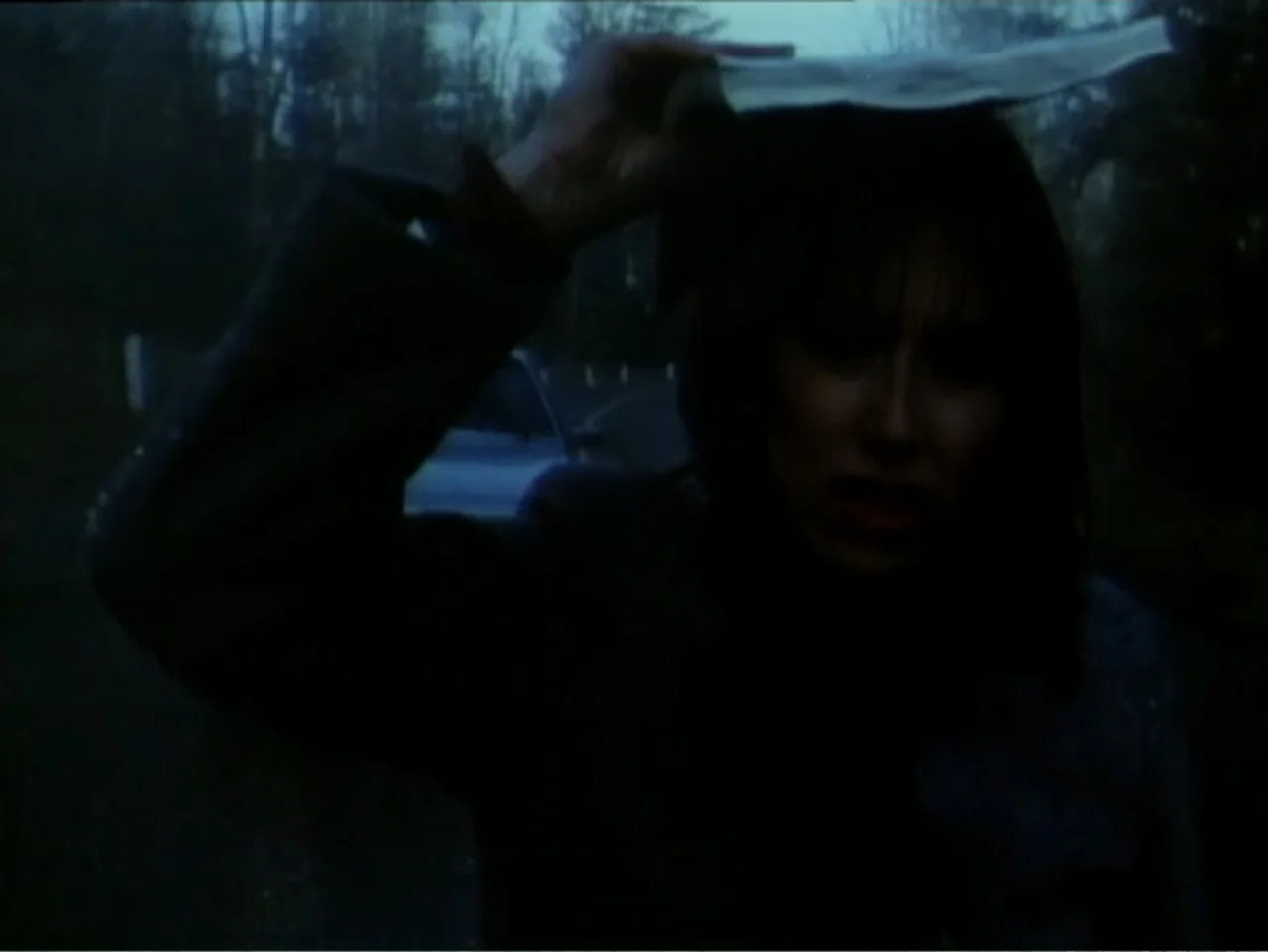 Curtains (1995) movie still: medium — A woman stands in a dark, outdoor location at night with a blurred vehicle behind. She holds a white piece of…