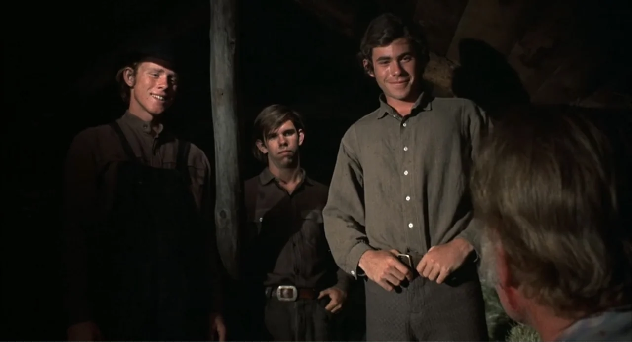 The Spikes Gang (1974) movie still: medium — A dark wooden shed interior with rough-hewn timbers visible in the background. Three young men stand…