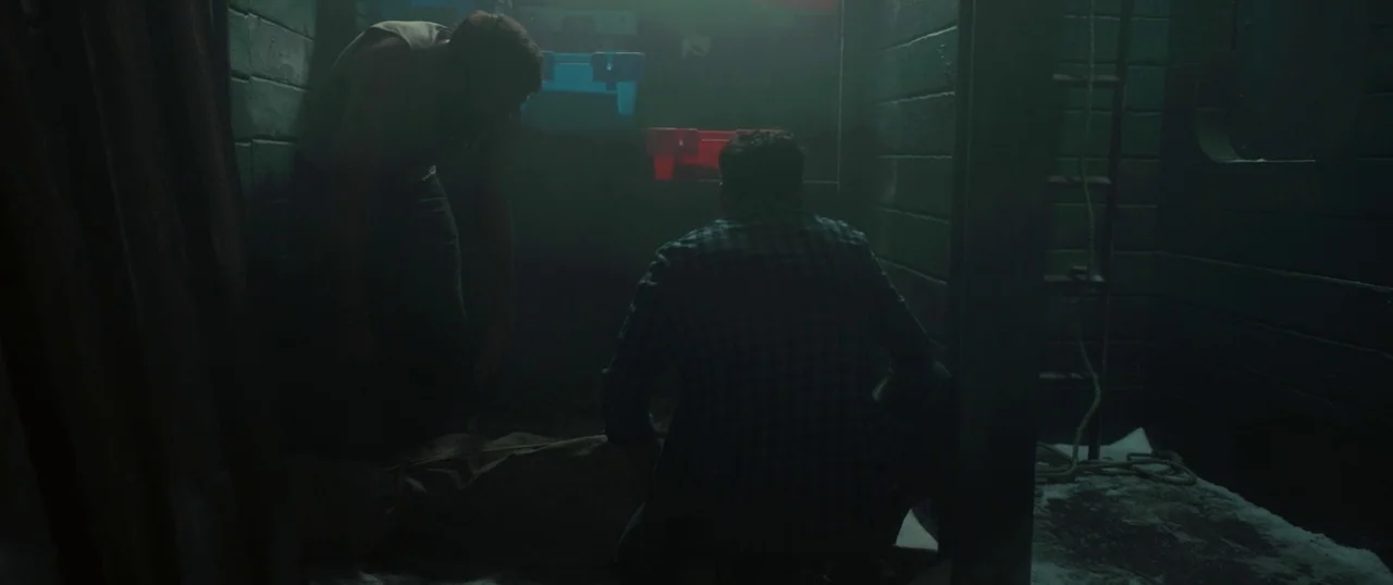 Sea Fever (2019) movie still: medium — A dimly lit concrete storage area with storage crates stacked on shelves. Two men kneel in the center,…