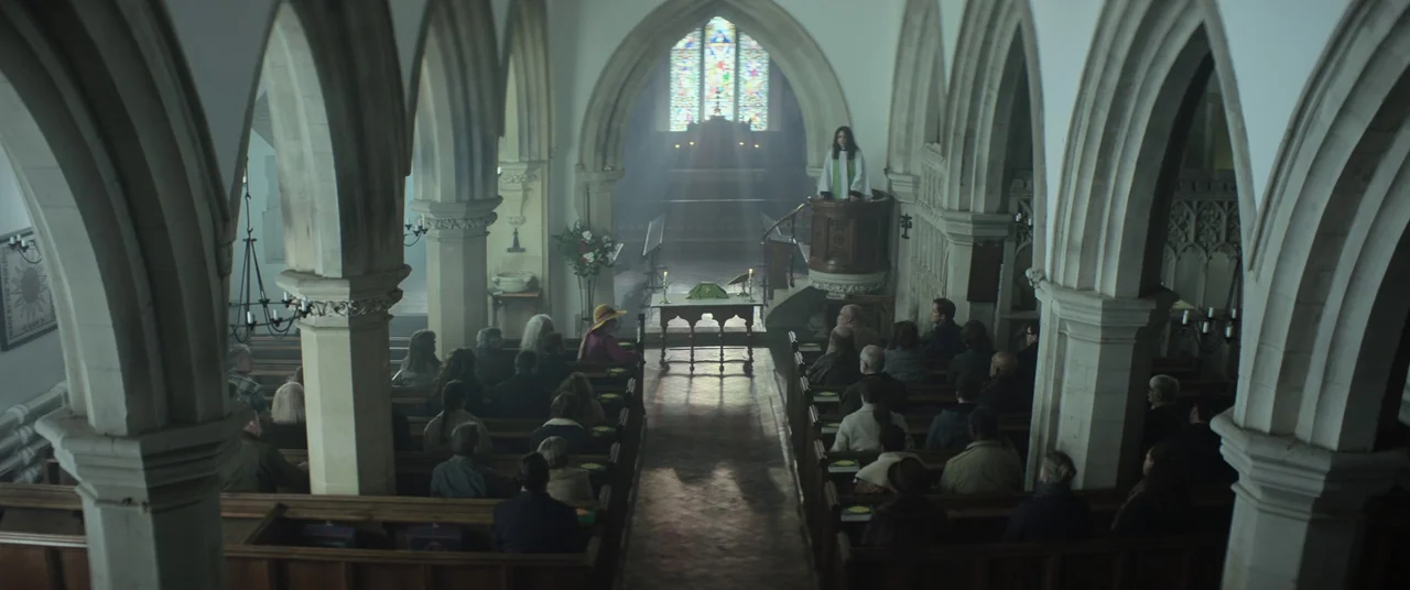 Lord of Misrule (2023) movie still: wide — A stone church nave features arched pillars and wooden pews filled with seated congregants. A person in…