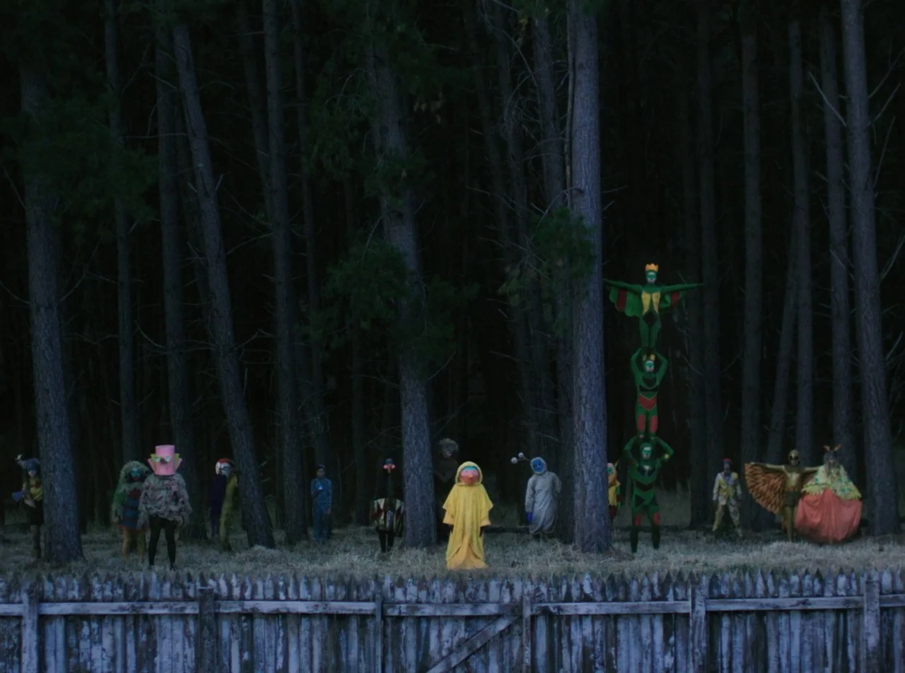 Girl Asleep (2015) movie still: wide — A group of people wearing eccentric creature costumes stand before a tall pine tree line. Three individuals…
